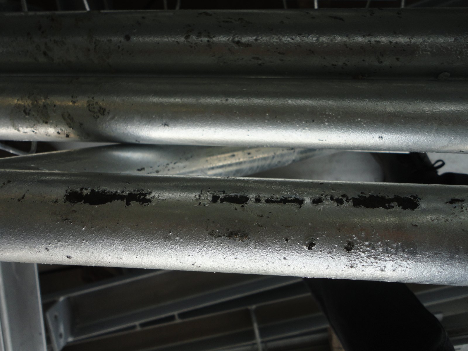 Galvanized steel tubes stacked on a rack: elongated black bare patches run along the upper face of the nearest tube where the zinc never took, with sound bright coating surrounding each patch