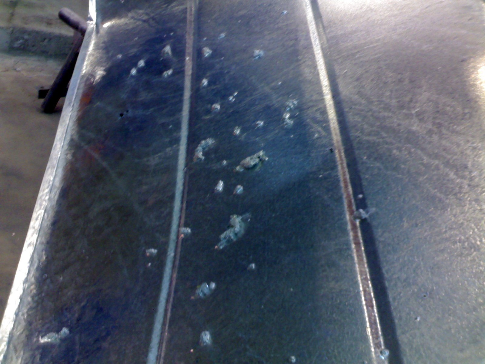 Wide galvanized panel with scattered raised blisters erupting through the coating, their pale broken crowns standing out against the smooth darker zinc surface