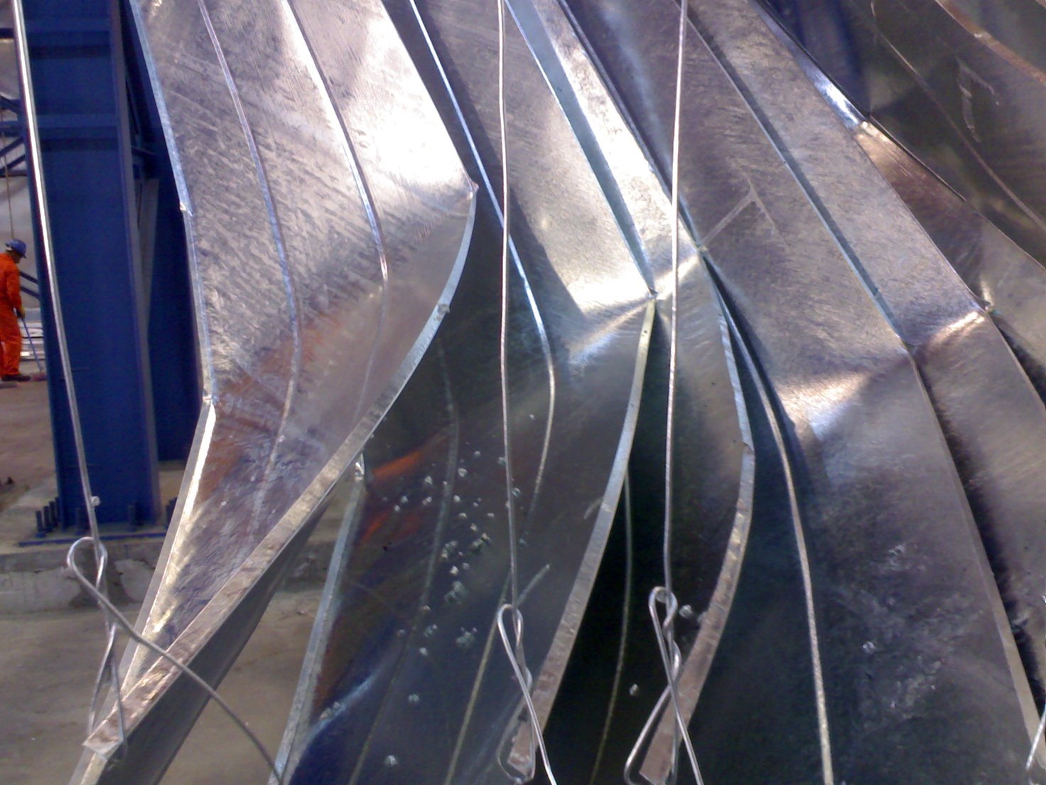 Thin galvanized steel plates hanging on wires after the kettle, buckled into deep S-curves and waves along their length while the zinc coating on them stays bright and sound