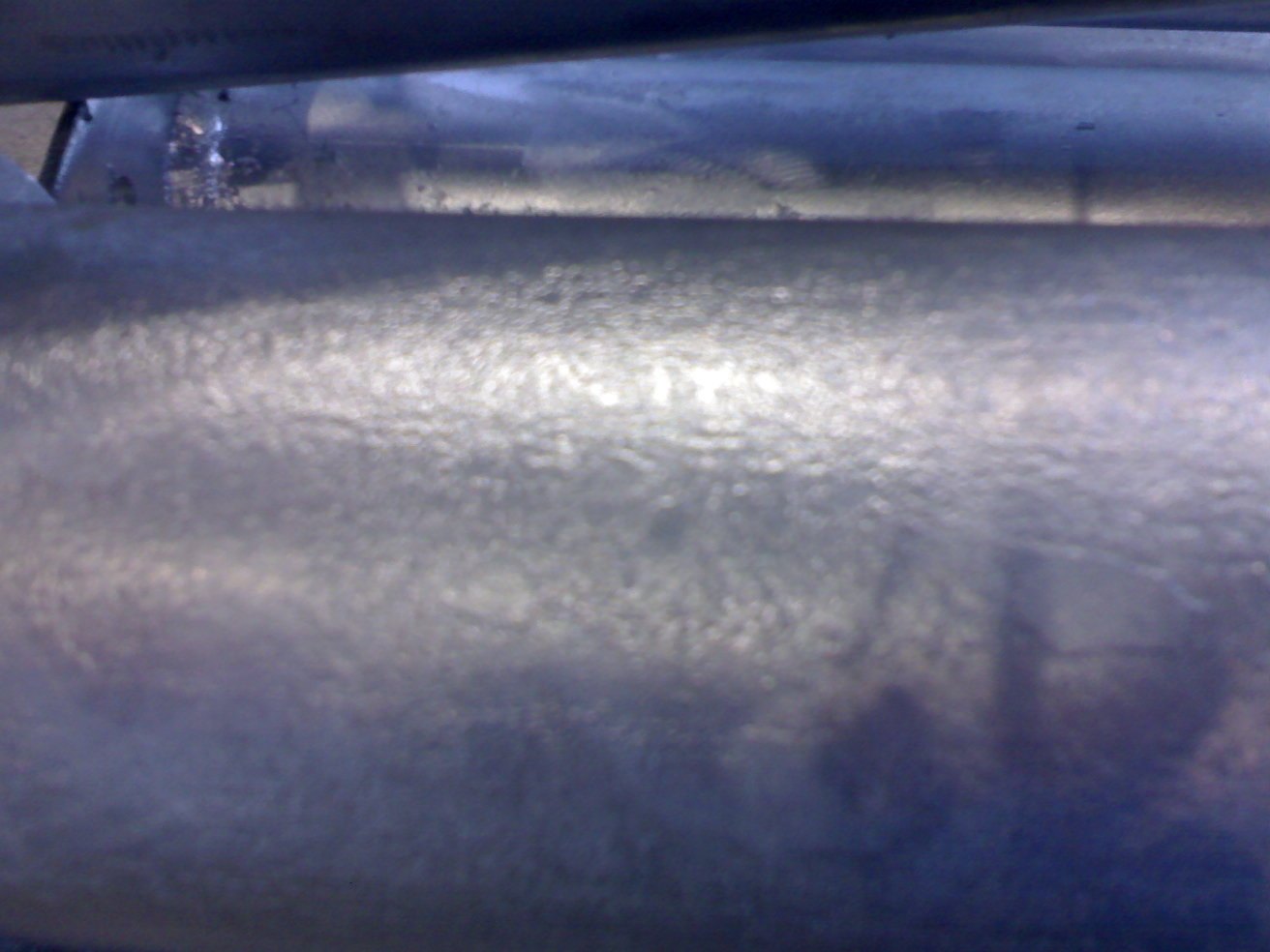 Galvanized tubular section photographed close up: a coarse granular coating texture shows along the band of raking light, with no isolated lumps or inclusions