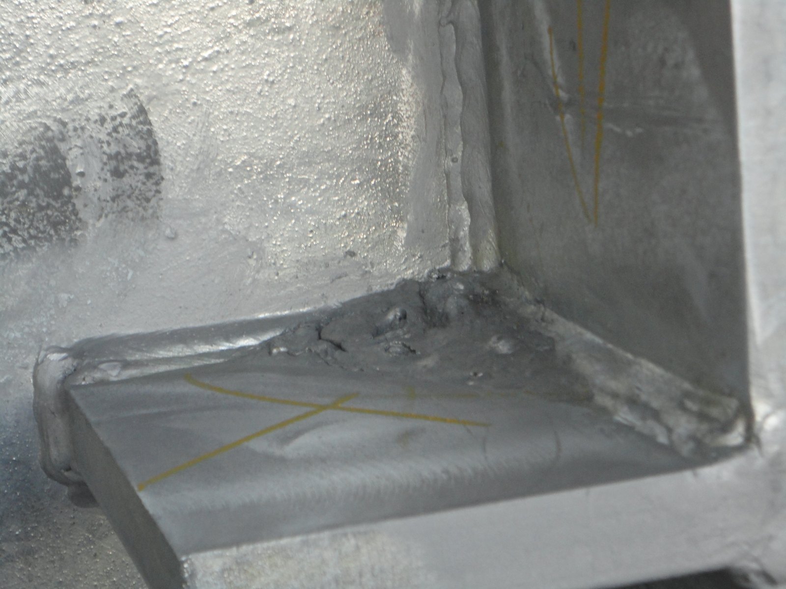 Internal corner of a galvanized steel fabrication: a thick lumpy mass of solidified zinc has pooled on the horizontal gusset plate, which has no drain hole, while the surrounding faces carry bright normal coating