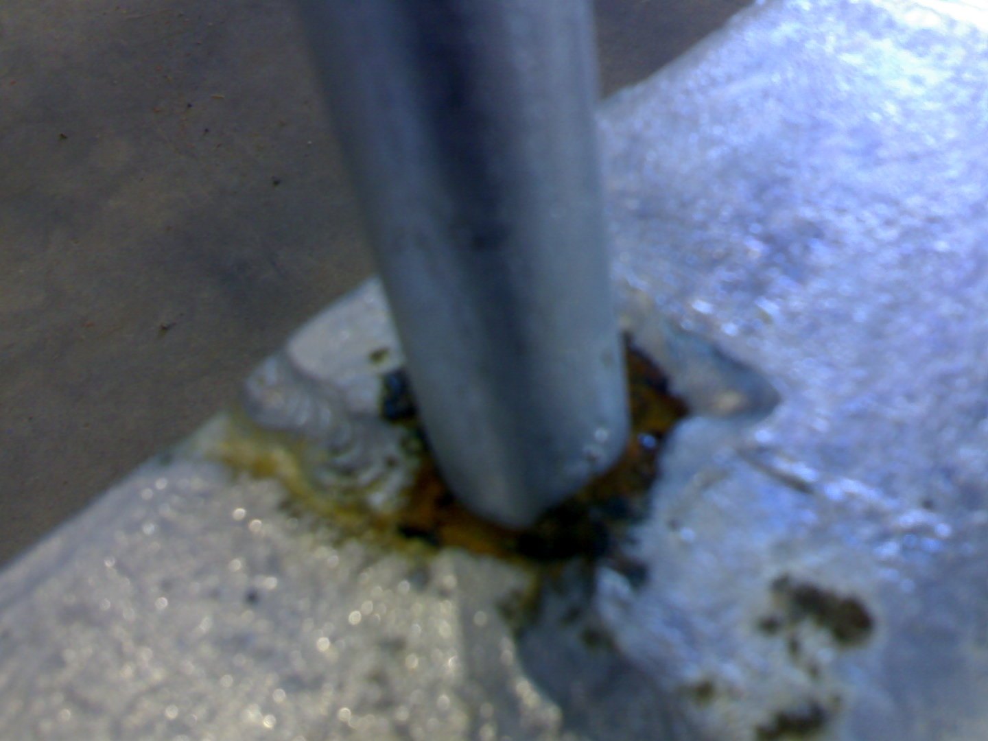 Galvanized bar welded into a base plate: translucent amber liquor weeps from the joint crevice and spreads over the surrounding sound coating, with darker crusted residue at the gap itself